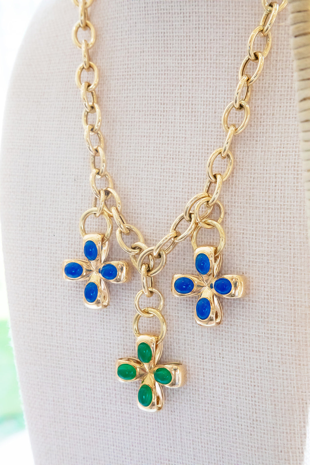 Gold-tone chain necklace with three dangling flower charms, two with blue stones and one with green stones, displayed on a cream fabric jewelry bust.  Made in Palm Beach Florida by local design team Pelimingo Palm Beach.  Clovers / Pendants/ crosses