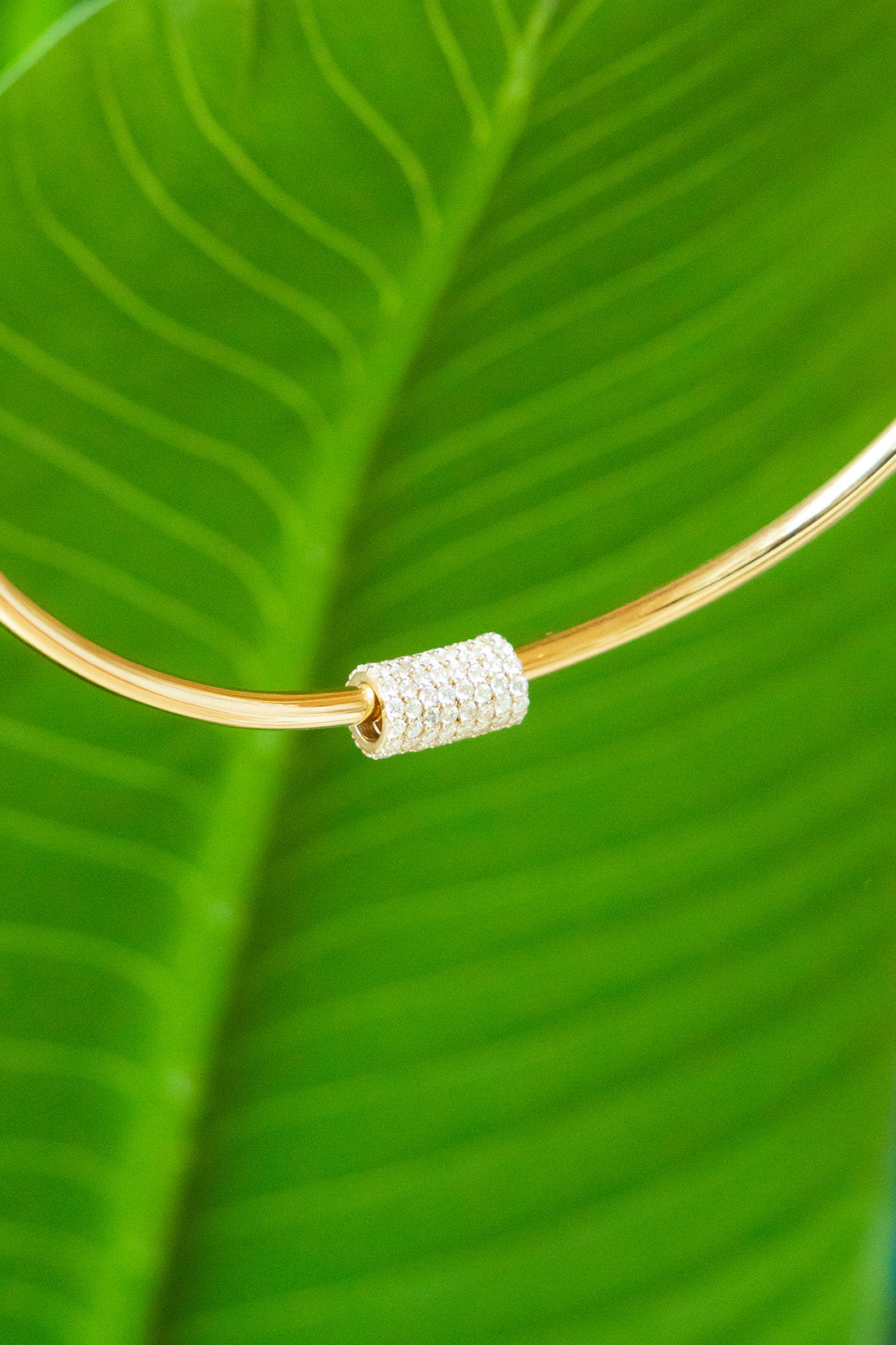 A close-up of a gold bangle bracelet featuring a textured, diamond-like silver cylindrical bead, set against a vibrant green tropical leaf background. Palm Beach Jewlery Designer, Pelimingo Palm Beach