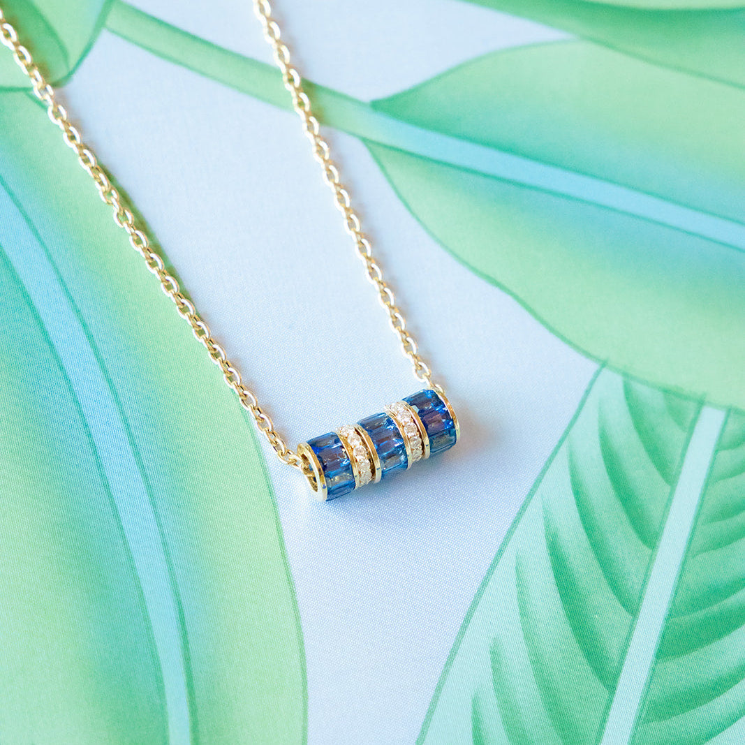 A gold chain necklace with a cylindrical pendant featuring blue and gold striped details, displayed on a tropical leaf print background. Palm Beach jewelry designers, Pelimingo Palm Beach