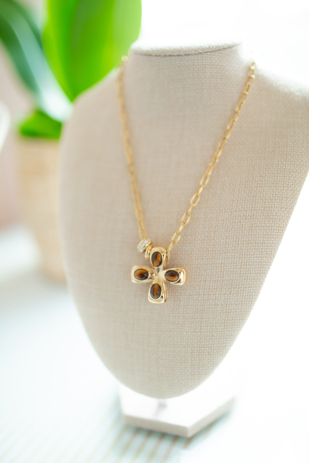 Gold necklace with a cross pendant on a white stand with a blurred green plant in the background. Tiger Eye Cabochon cross in 14K gold.  Palm Beach based Jewelry designers