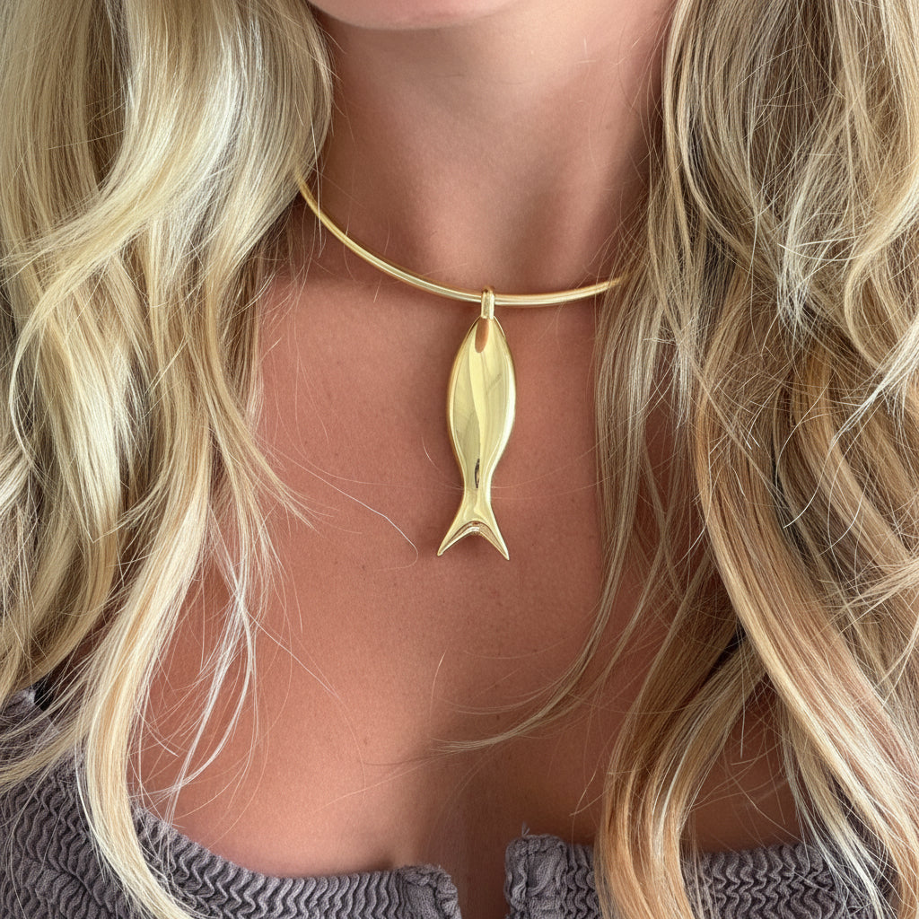 A close-up of a model's neck and chest wearing a delicate gold wire choker necklace with a polished gold fish pendant.