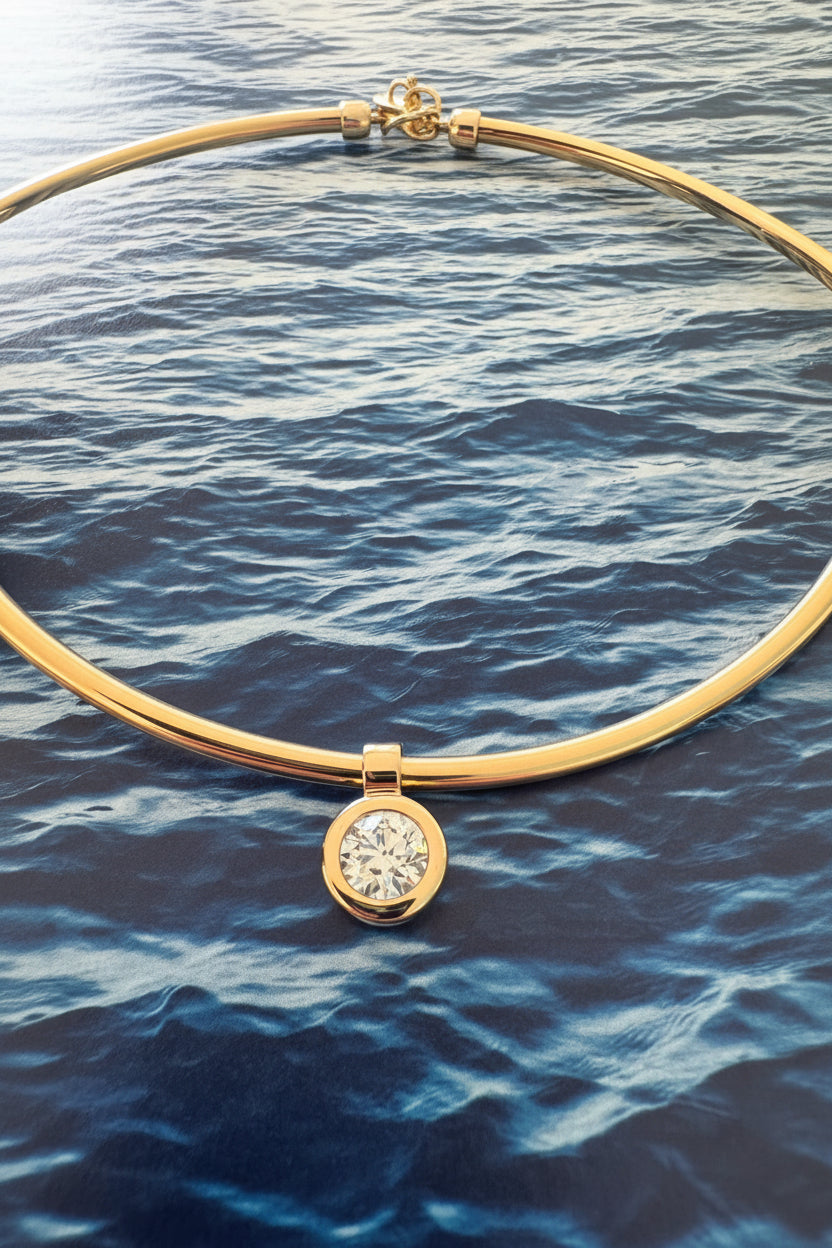 A gold-toned bangle necklace with a round bezel-set clear gemstone pendant, displayed against a printed background of blue ocean waves.