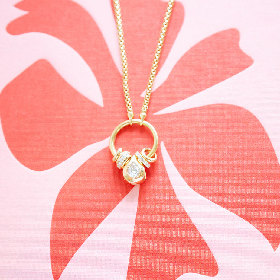 A gold necklace with a small ring pendant and clear gemstone charm, displayed on a pink card with a coral hibiscus flower graphic. The brand name 'ASSOULINE' is printed at the bottom.