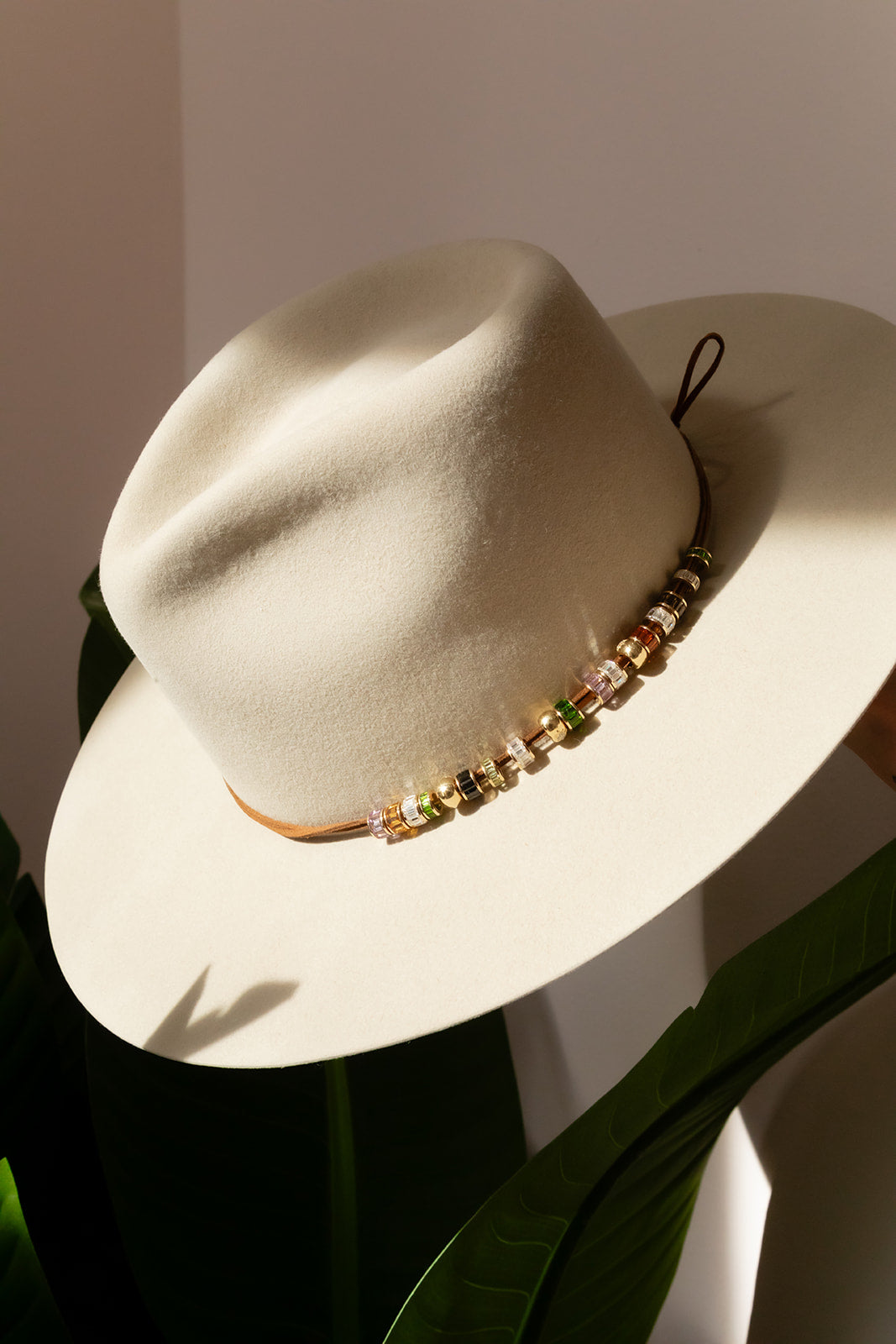 A cream-colored felt fedora hat with a colorful beaded band, styled with green leaves against a warm beige background.