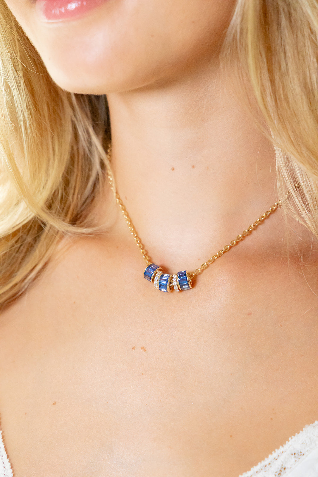 A close-up of a model wearing a delicate gold chain necklace with three small cylindrical charms featuring blue and gold striped patterns.
