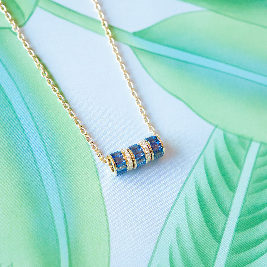 A gold chain necklace with a cylindrical pendant featuring blue and gold striped details, displayed on a tropical leaf print background.