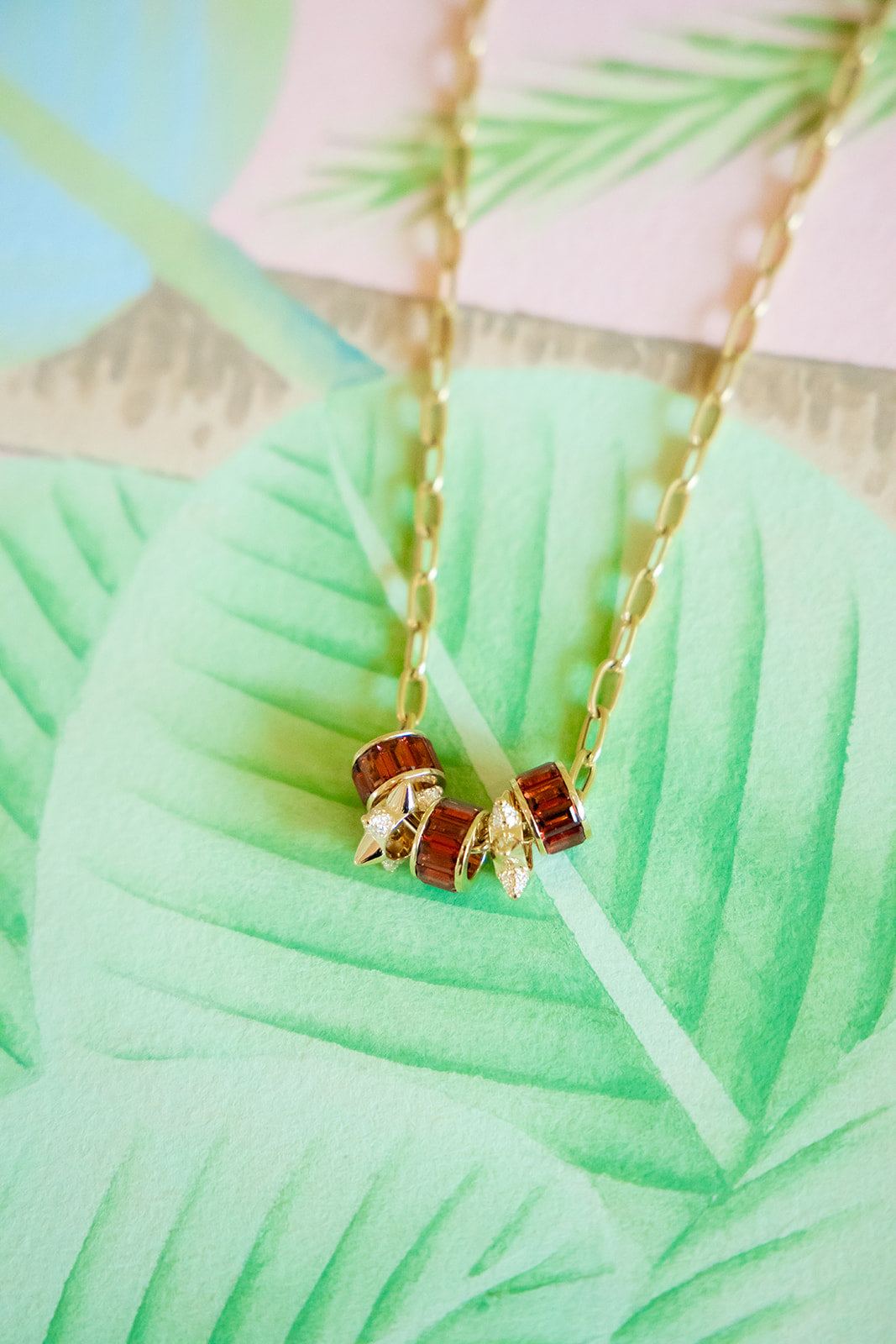 A gold chain necklace with three small cylindrical beads, two in reddish-brown and one in gold, displayed on a large mint green tropical leaf print background.