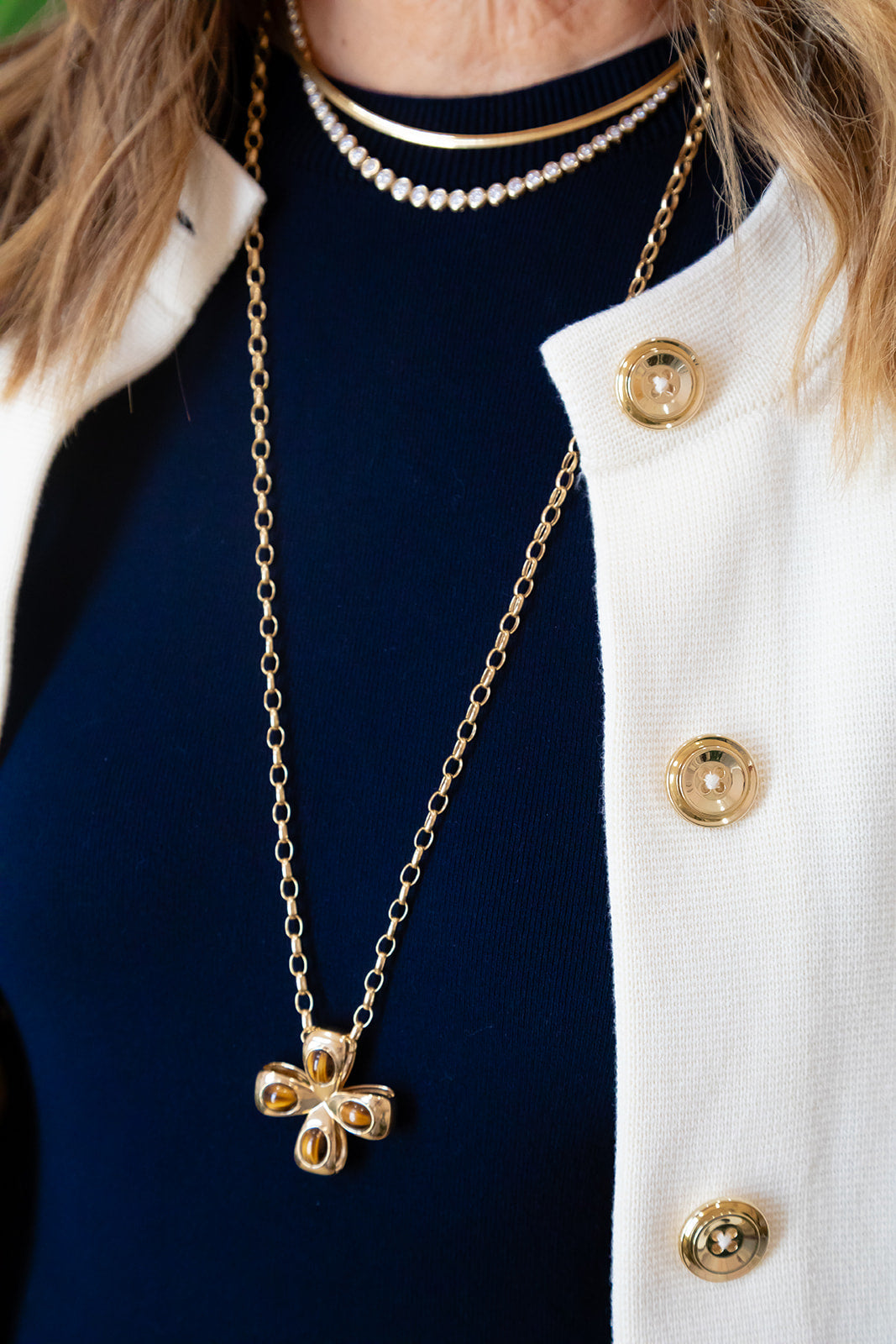 A close-up of a person wearing layered necklaces, including a gold clover pendant necklace over a dark navy top and white jacket. Chic cross or pendant in Tiger Eye and 14K gold.  Palm Beach Designers Pelimingo